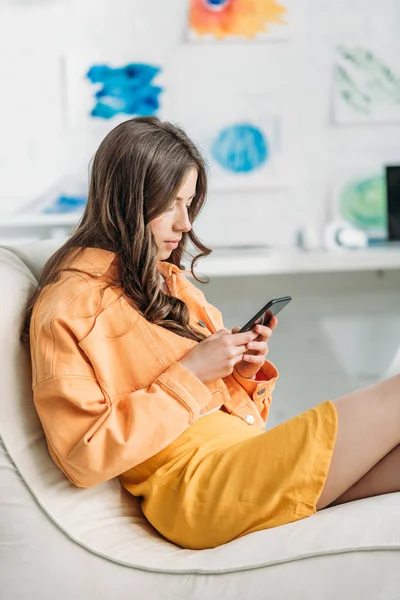 Jeune femme concentrée en fermeture orange à l'aide d'un smartphone à la maison — Photo de stock