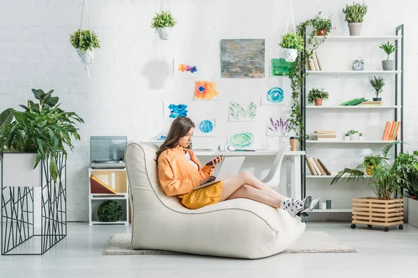 Young woman using laptop and smartphone while sitting on soft chaise lounge in spacious room — Stock Photo