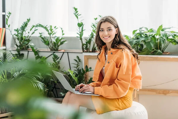 Foco seletivo da mulher alegre cercada por plantas verdes sorrindo ao usar laptop em casa — Fotografia de Stock