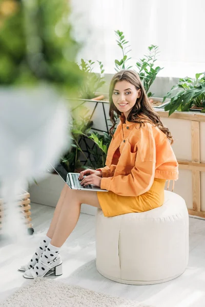 Selektiver Fokus einer glücklichen jungen Frau, die auf einem Hocker sitzt und zu Hause Laptop benutzt — Stockfoto