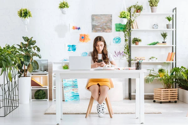 Junge Frau benutzt Smartphone, während sie mit Laptop am Schreibtisch in einem geräumigen Zimmer zu Hause sitzt — Stockfoto
