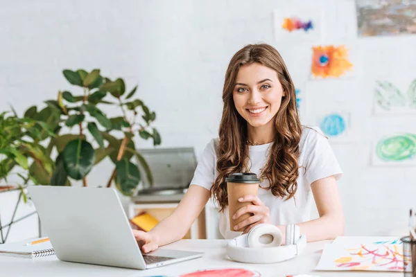 Sonriente mujer joven mirando a la cámara mientras usa el ordenador portátil y sostiene la taza desechable - foto de stock