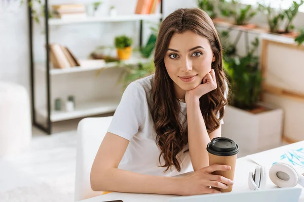 Sonriente joven mujer sosteniendo taza de papel y mirando a la cámara mientras está sentado en el escritorio en casa - foto de stock