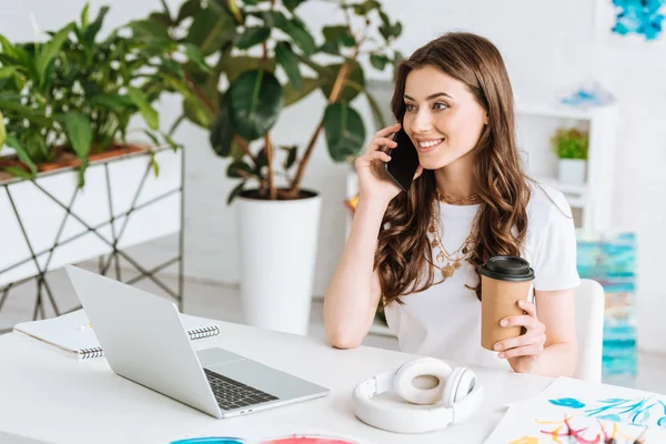 Chica sonriente hablando en el teléfono inteligente y sosteniendo la taza de papel mientras está sentado en la mesa con el ordenador portátil y auriculares - foto de stock