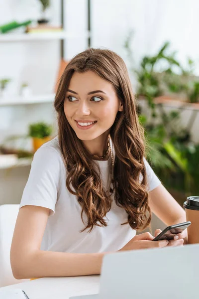 Sorrindo jovem mulher olhando para longe enquanto sentado na mesa e usando smartphone — Fotografia de Stock