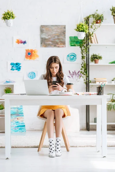 Young woman using smartphone while sitting at desk in spacious room with paintings on wall — Stock Photo