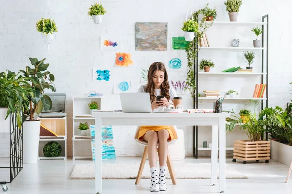 Bella giovane donna utilizzando smartphone mentre seduto in camera spaziosa con piante verdi e dipinti a parete — Foto stock