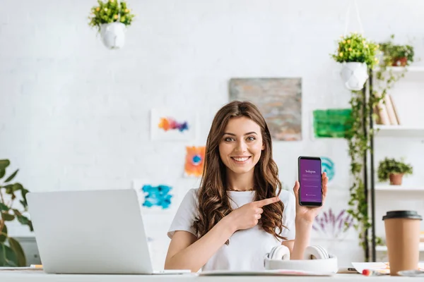 KYIV, UKRAINE - APRIL 17, 2019: Cheerful young woman pointing with finger at smartphone with Instagram app on screen. — Stock Photo