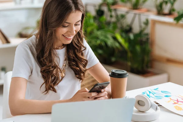 Mujer hermosa feliz usando el ordenador portátil mientras está sentado en el escritorio cerca de la computadora portátil y taza de papel - foto de stock