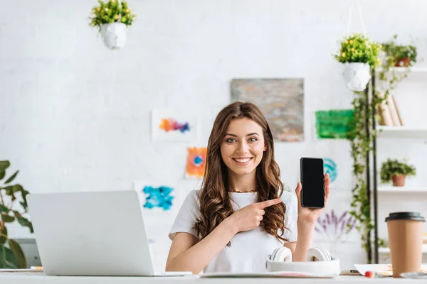 Belle fille regardant la caméra et pointant vers le smartphone avec écran vide . — Photo de stock