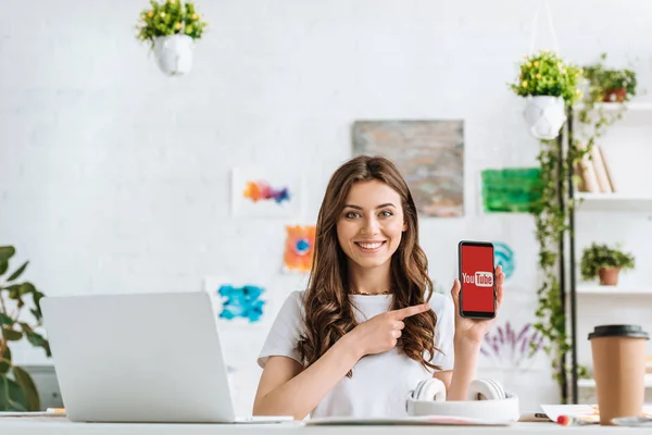 KYIV, UKRAINE - APRIL 17, 2019: Cheerful young woman looking at camera and pointing with finger at smartphone with Skype app on screen. — Stock Photo