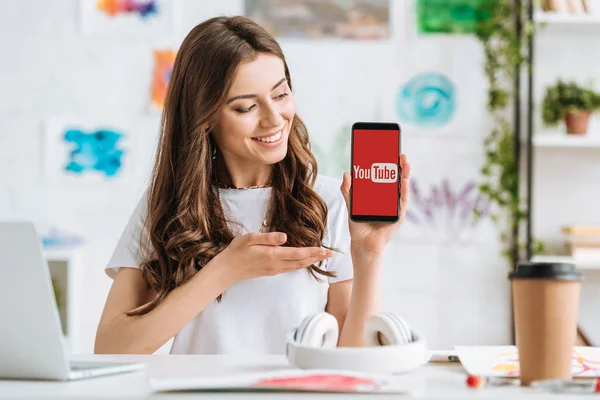 KYIV, UKRAINE - APRIL 17, 2019: Happy young woman pointing with hand at smartphone with Youtube app on screen. — Stock Photo