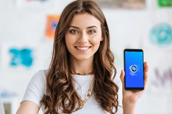 KYIV, UCRANIA - 17 de abril de 2019: Hermosa chica mirando a la cámara y mostrando el teléfono inteligente con la aplicación Shazam en la pantalla . - foto de stock