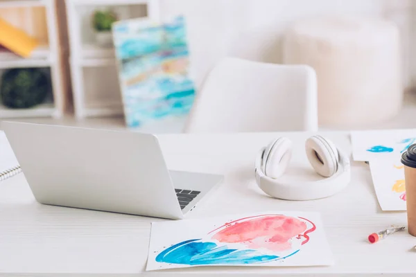 Laptop, headphones and paintings on white tabletop near colorful paitings — Stock Photo