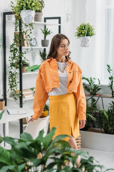 Селективное внимание задумчивой молодой женщины, стоящей рядом со стулом в просторной комнате, украшенной зелеными растениями — стоковое фото