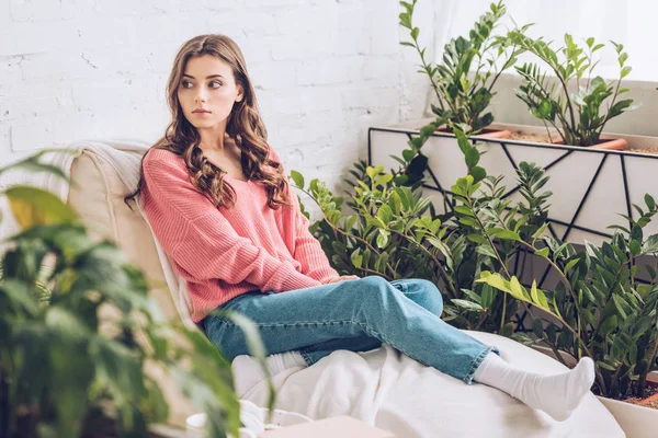 Селективное внимание задумчивой молодой женщины, сидящей в комнате рядом с пышными зелеными растениями — стоковое фото