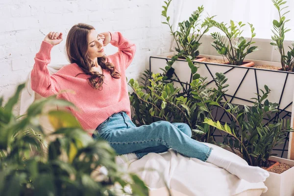 Selektiver Fokus einer hübschen jungen Frau, die sich dehnt, während sie im Raum in der Nähe üppiger grüner Pflanzen sitzt — Stockfoto