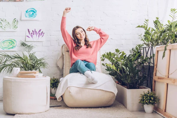 Smiling girl stretching while sitting on soft chaise lounge near pouf and lush green plants — Stock Photo