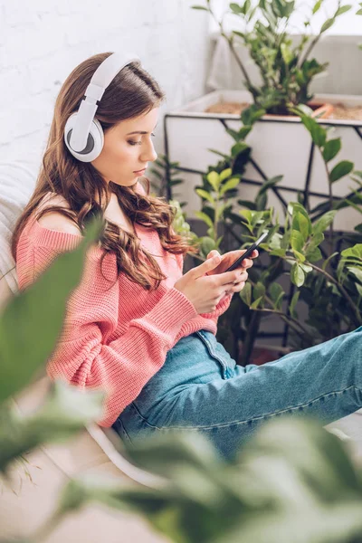Selective focus of pensive girl listening music in headphones and using smartphone while sitting surrounded by green plants — Stock Photo