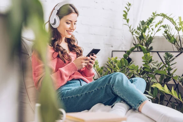 Foco seletivo de menina sorridente usando smartphone enquanto sentado cercado por plantas verdes e ouvir música em fones de ouvido — Fotografia de Stock