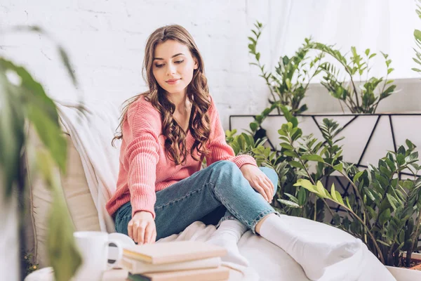Selektiver Fokus eines attraktiven Mädchens, das ein Buch nimmt, während es zu Hause in einer weißen Chaiselongue sitzt — Stockfoto