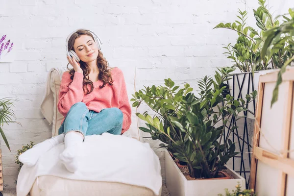 Pretty girl with closed eyes relaxing while sitting in soft chaise lounge and listening music in headphones — Stock Photo