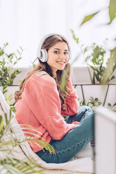 Atractiva joven escuchando música en auriculares y mirando a la cámara mientras está sentada rodeada de plantas verdes en casa - foto de stock