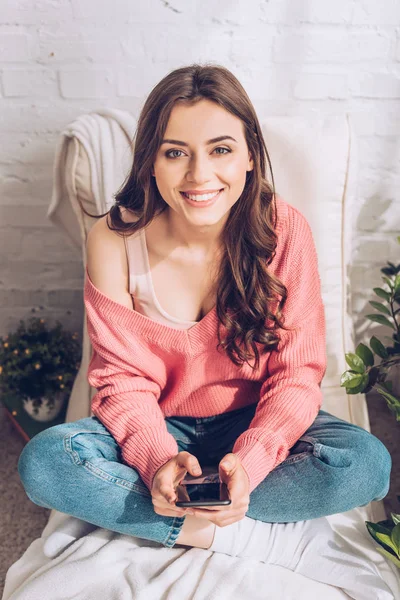 Happy beautiful girl smiling at camera while sitting with crossed legs and using smartphone — Stock Photo