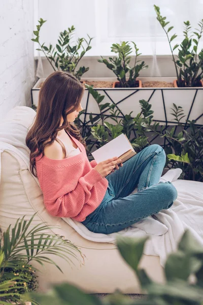 Foco seletivo de livro de leitura jovem mulher enquanto sentado com pernas cruzadas perto de plantas verdes em casa — Fotografia de Stock