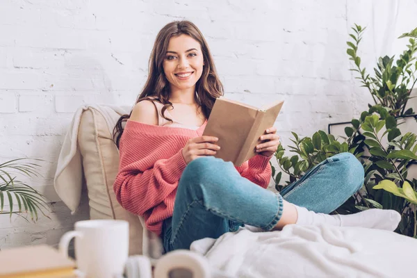 Attraktive fröhliche Mädchen lächelt in die Kamera, während sie mit Buch auf weicher Chaiselongue sitzt — Stockfoto