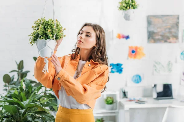 Beautiful woman in orange clothing touching hanging flowerpot at home — Stock Photo