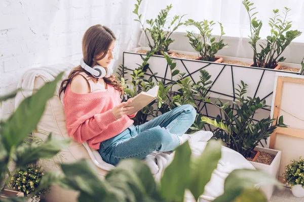 Selective focus of young woman reading book while sitting with crossed legs surrounded by lush plants at home — Stock Photo