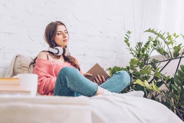 Mujer bonita pensativa mirando hacia otro lado mientras está sentado con las piernas cruzadas y sosteniendo libro - foto de stock