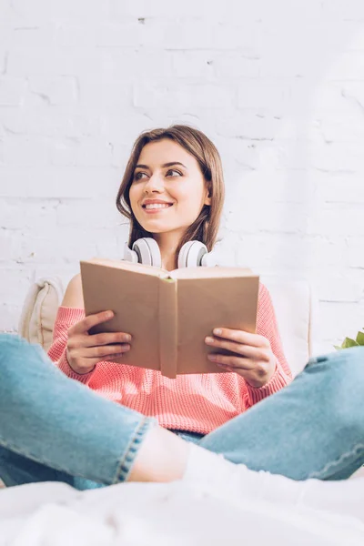 Femme souriante regardant loin tout en étant assis avec les jambes croisées et tenant le livre — Photo de stock