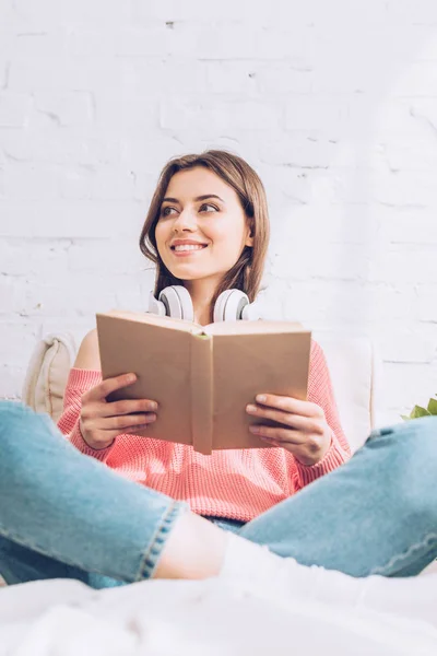 Donna sorridente che distoglie lo sguardo mentre siede con le gambe incrociate e tiene il libro — Foto stock