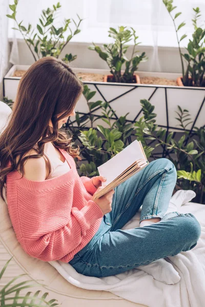 Junge Frau liest Buch, während sie zu Hause von grünen Pflanzen umgeben ist — Stockfoto