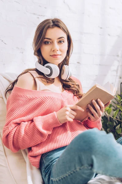 Attrayant jeune femme avec livre et écouteurs regardant la caméra tout en étant assis avec les jambes croisées — Photo de stock