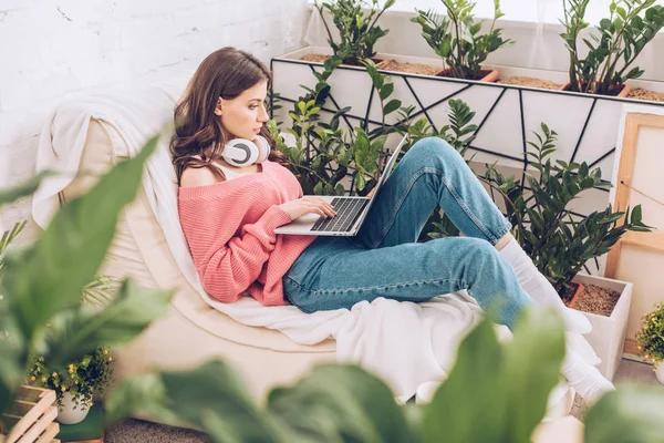 Вибірковий фокус красивої дівчини з навушниками на шиї за допомогою ноутбука, сидячи на м'якому шезлозі — стокове фото
