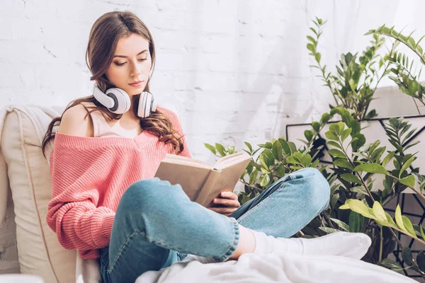 Mulher atenta com fones de ouvido no pescoço leitura livro enquanto sentado com pernas cruzadas — Fotografia de Stock