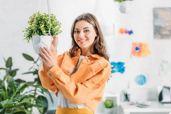 Cheerful woman touching hanging flowerpot with green plant and smiling at camera — Stock Photo