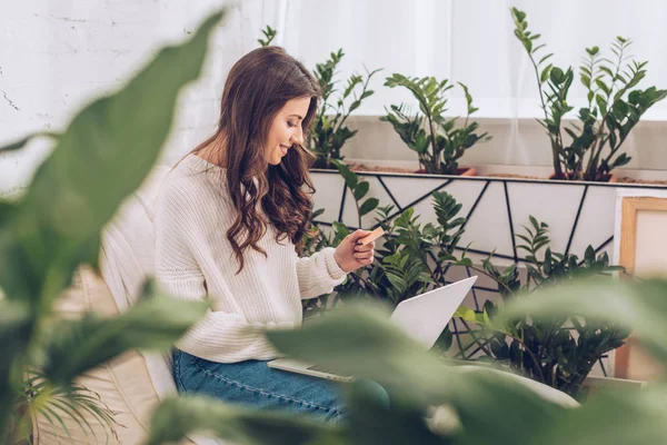 Foyer sélectif de la femme souriante tenant la carte de crédit et en utilisant un ordinateur portable tout en étant assis entouré de plantes vertes à la maison — Photo de stock