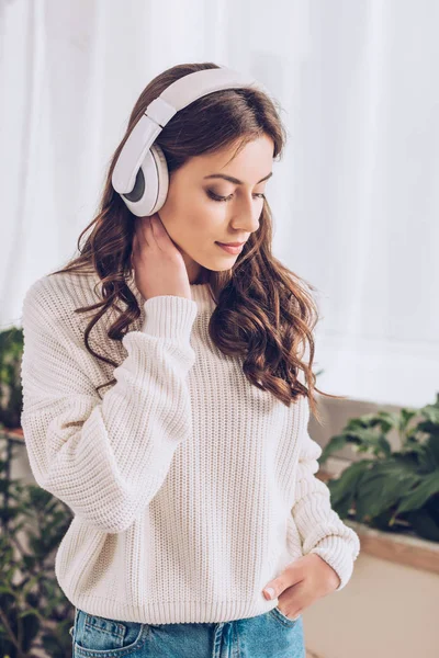 Pensive young girl listening music in headphones, holding hand in pocket and looking down — Stock Photo