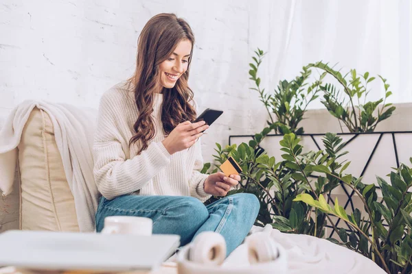 Selektiver Fokus einer hübschen jungen Frau, die ihr Smartphone nutzt, während sie zu Hause in der Nähe grüner Pflanzen sitzt — Stockfoto