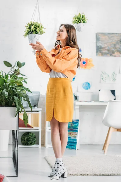 Trendy woman in orange clothing touching flowerpot with green plant — Stock Photo