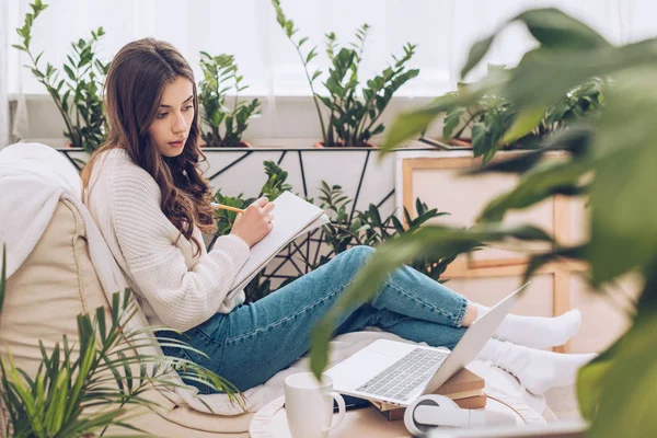 Foco seletivo da jovem atenta escrita usando laptop e escrita em notebook enquanto sentado perto de plantas verdes em casa — Fotografia de Stock