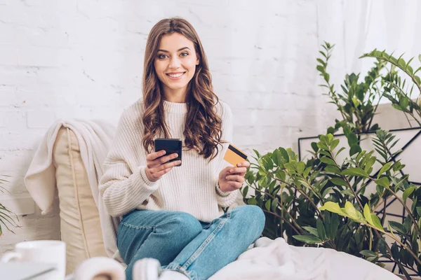 Feliz chica bonita sosteniendo la tarjeta de crédito y el uso de teléfono inteligente mientras sonríe a la cámara - foto de stock