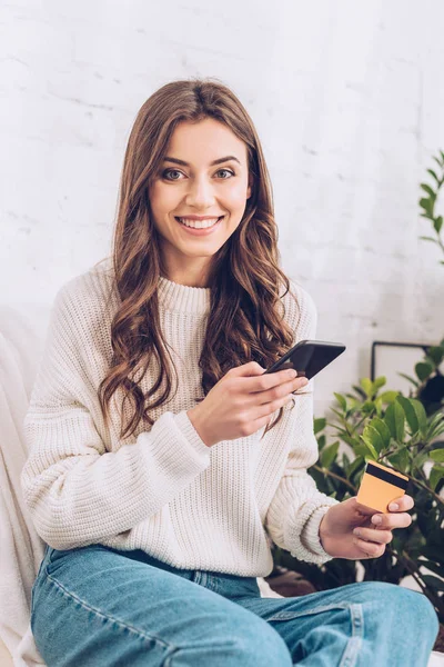 Счастливая молодая женщина смотрит в камеру, держа кредитную карту и смартфон — стоковое фото