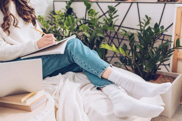 Cropped view of young woman writing in notebook while sitting near green plants at home — Stock Photo