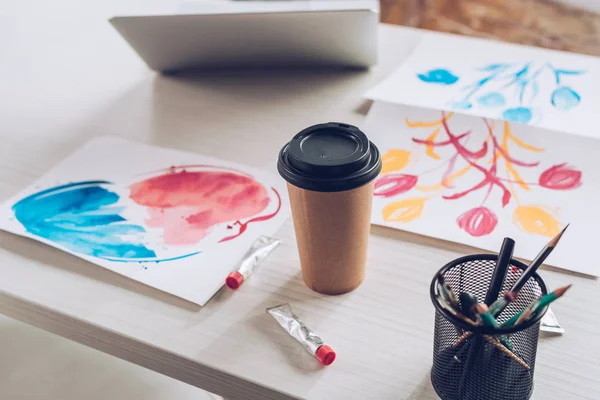 Tabletop with laptop, disposable cup, paint tubes, stationery and colorful paintings — Stock Photo