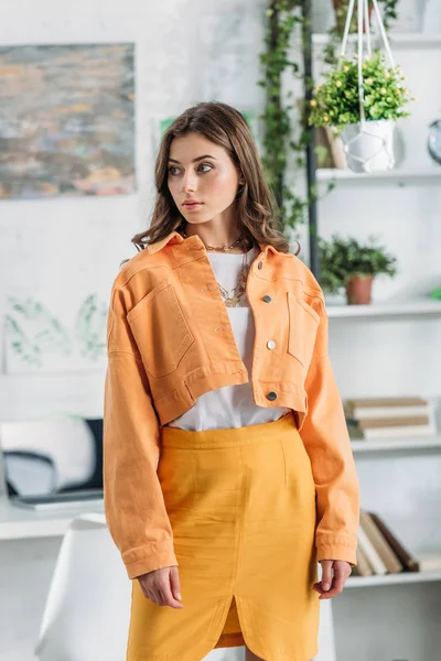 Задумчивая красивая девушка в оранжевой одежде смотрит в сторону, стоя рядом с стойкой с книгами и растениями — Stock Photo